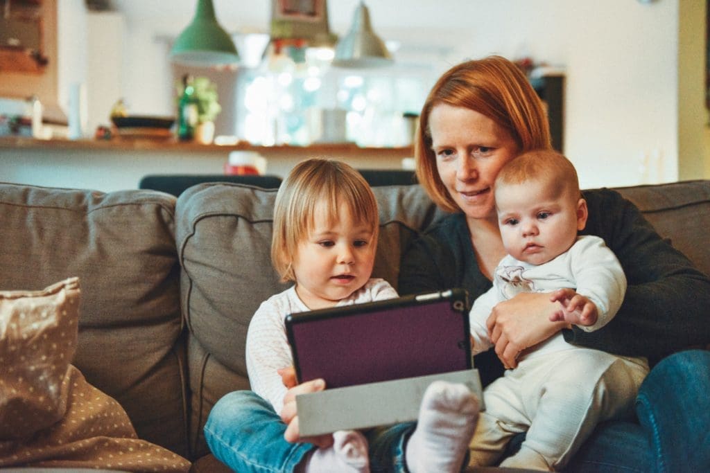 Woman with red hair holding her a toddler and baby looking at a tablet