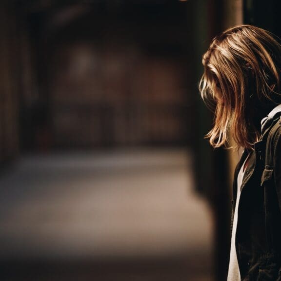 Image of a young woman taken from the side. She is looking at the floor and her hair is covering her face. She is standing alone in a dark outdoor space.