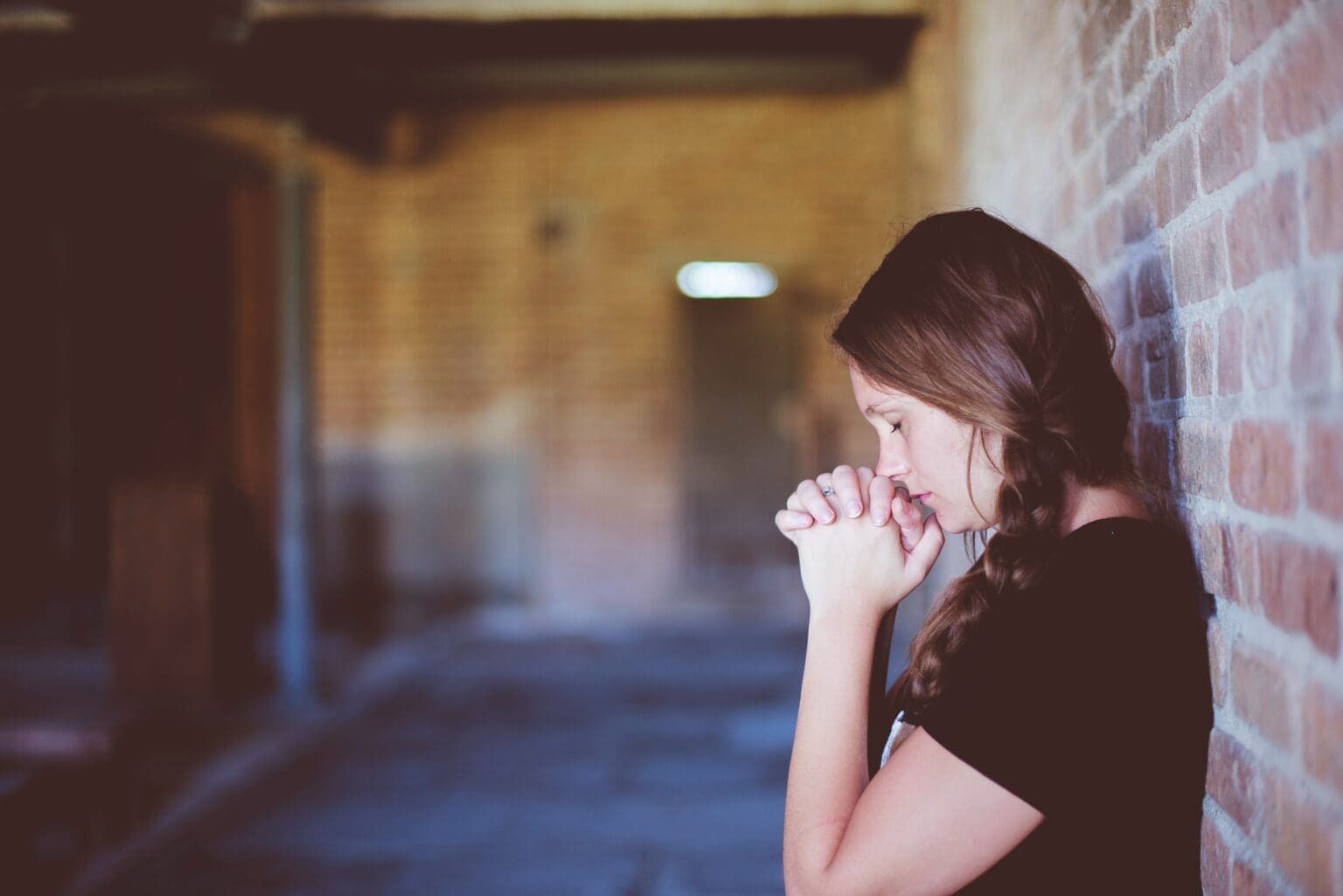Woman standing with her back against a wall. Her eyes are closed and she is holding her hands to her face in a prayer position.