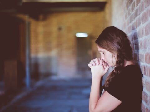 Woman standing with her back against a wall. Her eyes are closed and she is holding her hands to her face in a prayer position.