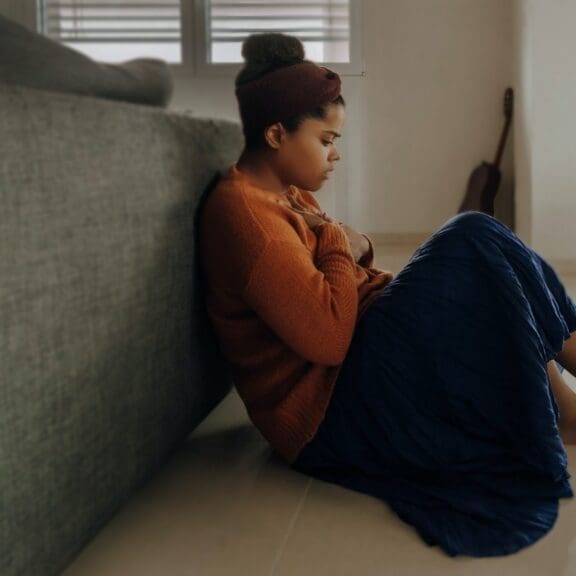 Woman sitting on the floor with her back against a sofa. She looks distressed.