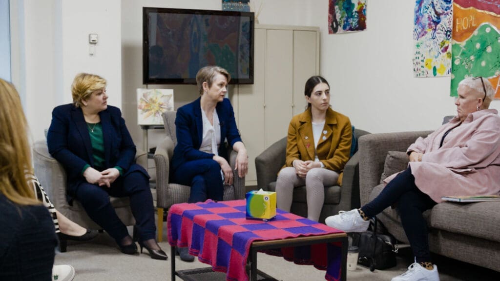 Four women sit on armchairs and a sofa facing and talking to each other, with a coffee table in front of them.