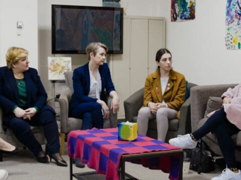 Four women sit on armchairs and a sofa facing and talking to each other, with a coffee table in front of them.