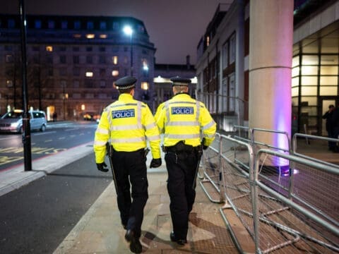 Two policemen in fluorescent uniform jackets pictured from behind walking down a street at night.
