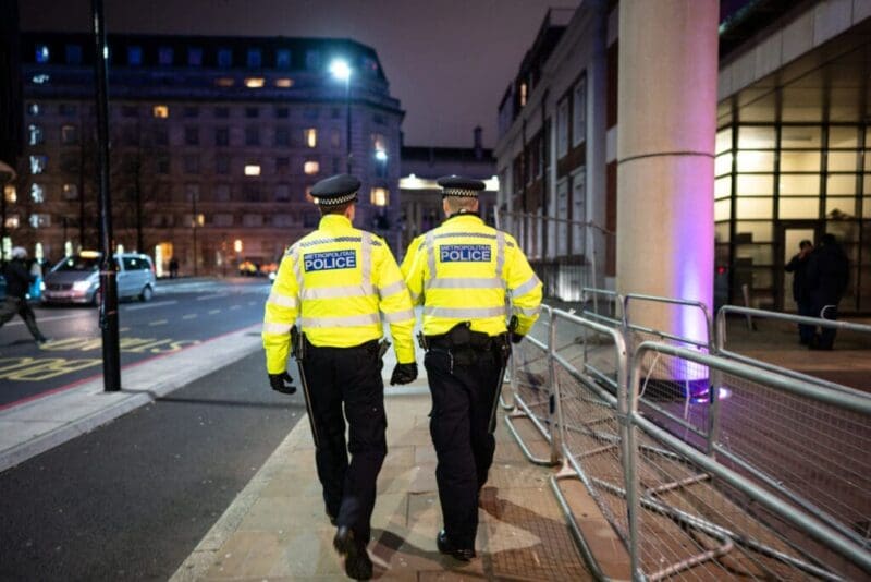 Two policemen in fluorescent uniform jackets pictured from behind walking down a street at night.