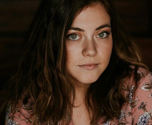 woman with brown curly hair wearing a floral top looking to camera with thoughtful expression