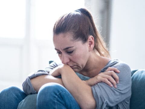 A woman with a ponytail sitting on a sofa slightly hunched over, with her knees raised to her chest and her chin and arms resting on her knees.