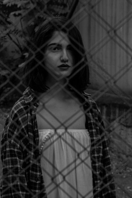 Sad faced woman with dark hair looking through a fence. She is wearing a white top and checked shirt