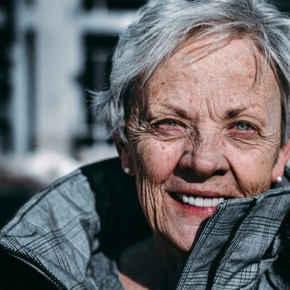 Pensioner-aged woman with short grey hair smiling at the camera, and stood outside a building in her coat with collar up