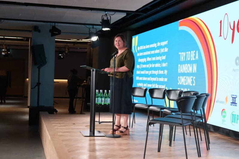 woman in dress stood on stage speaking in front of a screen behind displaying an image of the Angelou Partnership