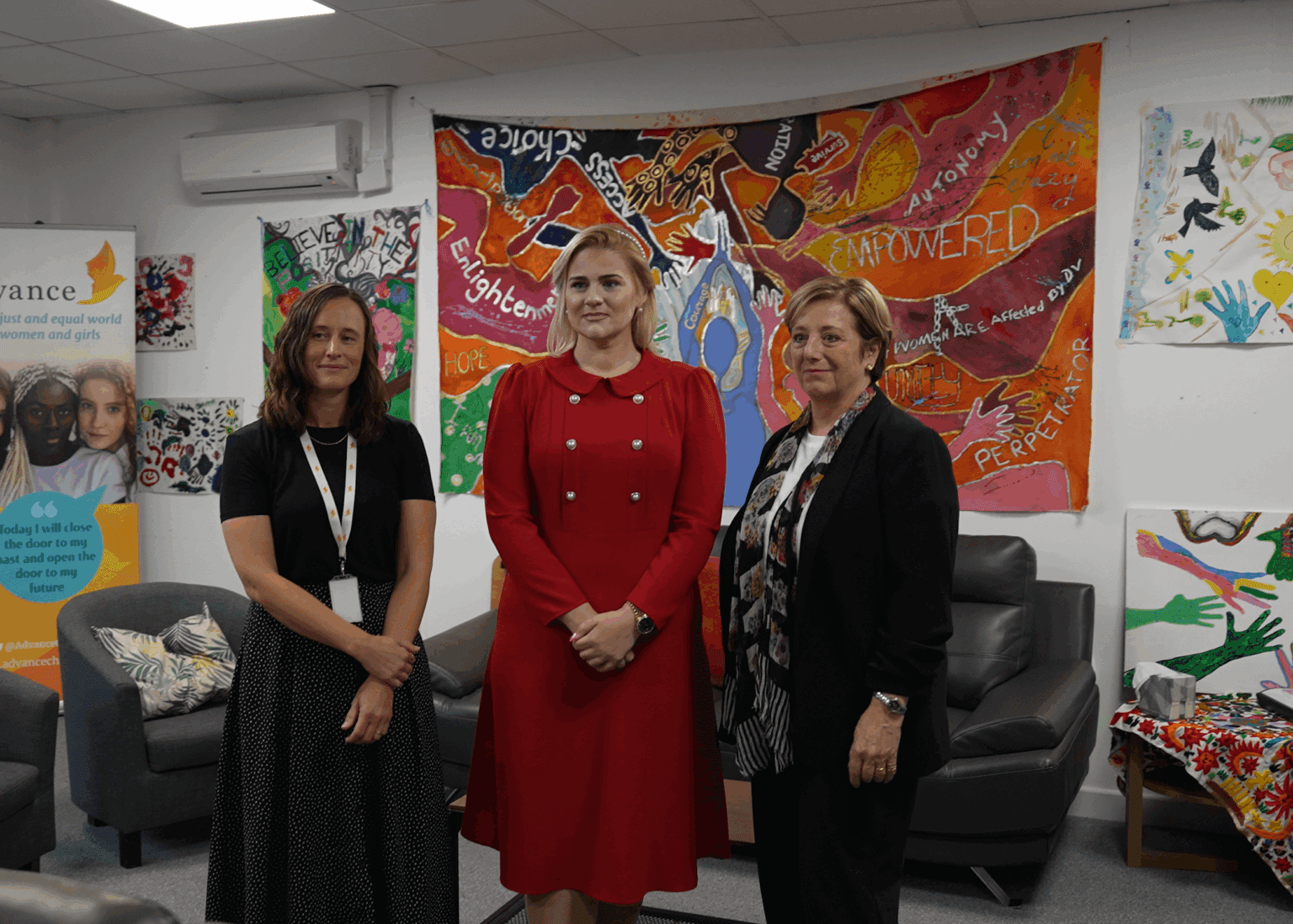 Three women stood beside each other in front of a colourful mural. Two are wearing black. One is wearing a red dress. They are all looking to camera and smiling.