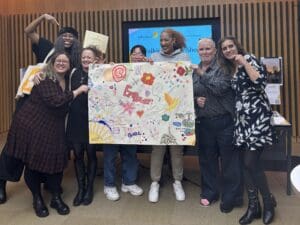 Group of women of varying ages holding up a mural they've created on white paper with colours pens and paints. Smiling at the camera and looking proud.