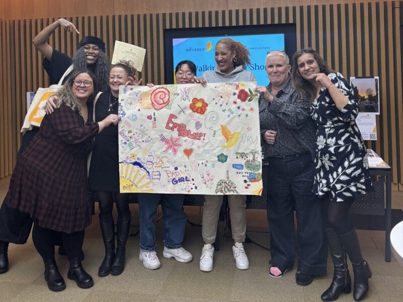 Group of women of varying ages holding up a mural they've created on white paper with colours pens and paints. Smiling at the camera and looking proud.