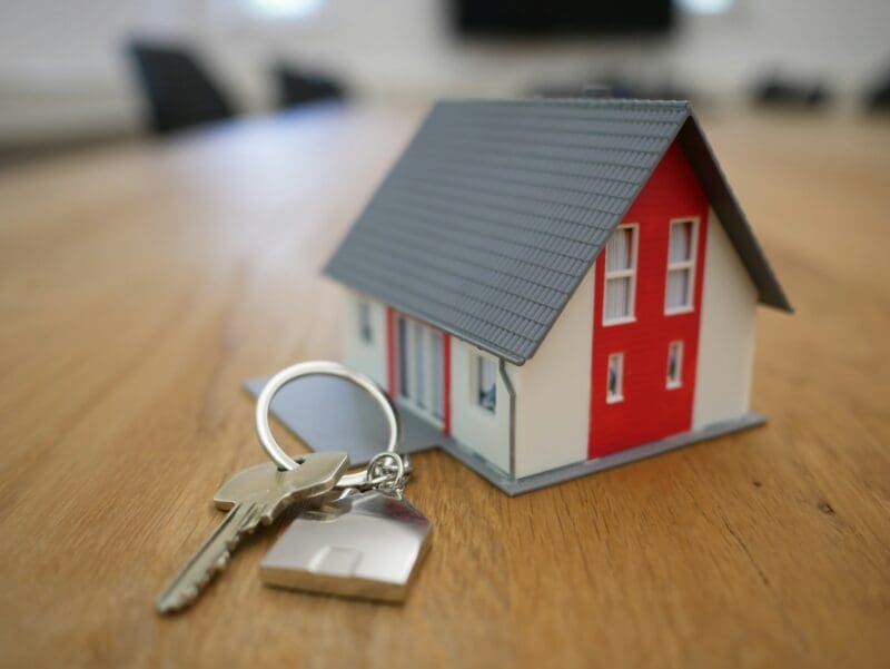 Small model house with grey roof and red door. On a pine table. Next to a house key on a keyring.