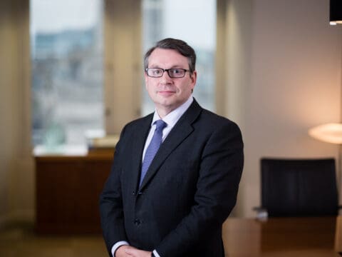 Man in his 40s with brown hair, wearing glasses. Dark suit jacket and a blue tie and white shirt. Smiling to camera. Hands clasped.