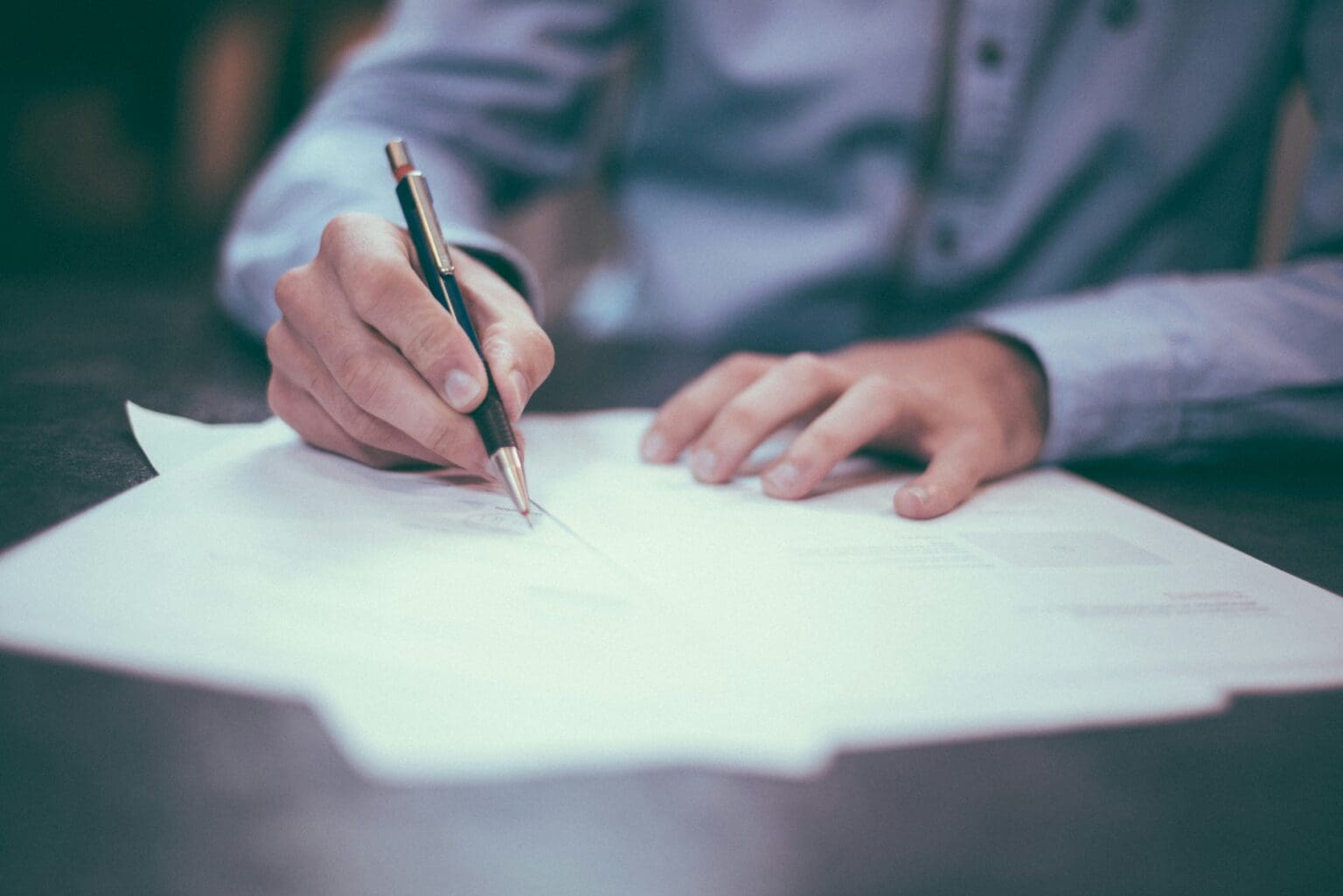someone sat at a desk, writing with a pen on paper. All you can see is the paper, pen person's hands and part of their body. They are wearing a blue shirt.