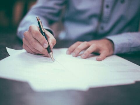 someone sat at a desk, writing with a pen on paper. All you can see is the paper, pen person's hands and part of their body. They are wearing a blue shirt.