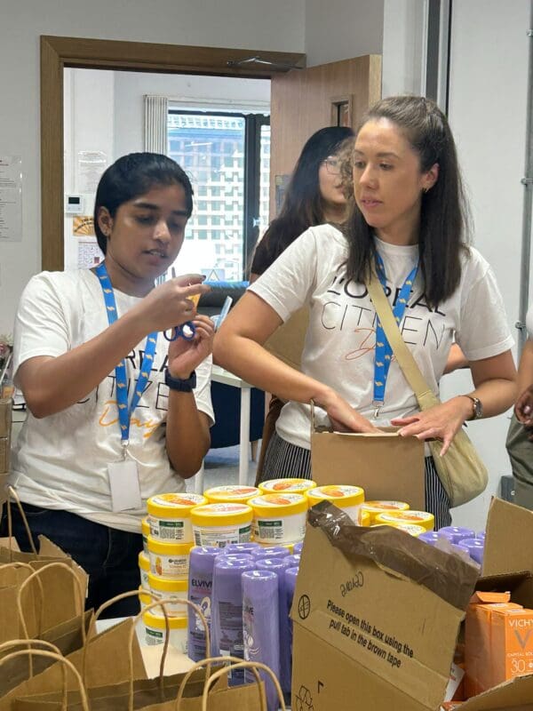 Two young women, one Asian, one caucasian, in white tshirts, standing at a table packing goodie bags with hair products and beauty products. Both are wearing lanyards