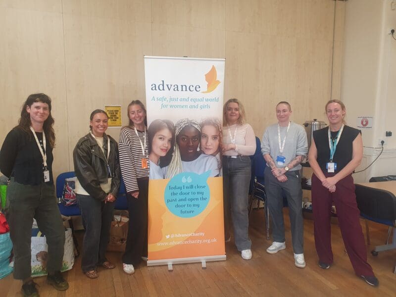 six women standing together in a line, flanking a banner promoting Advance. The women are smiling at the camera and are all dressed in trousers. Aged between early 20s to late 30.